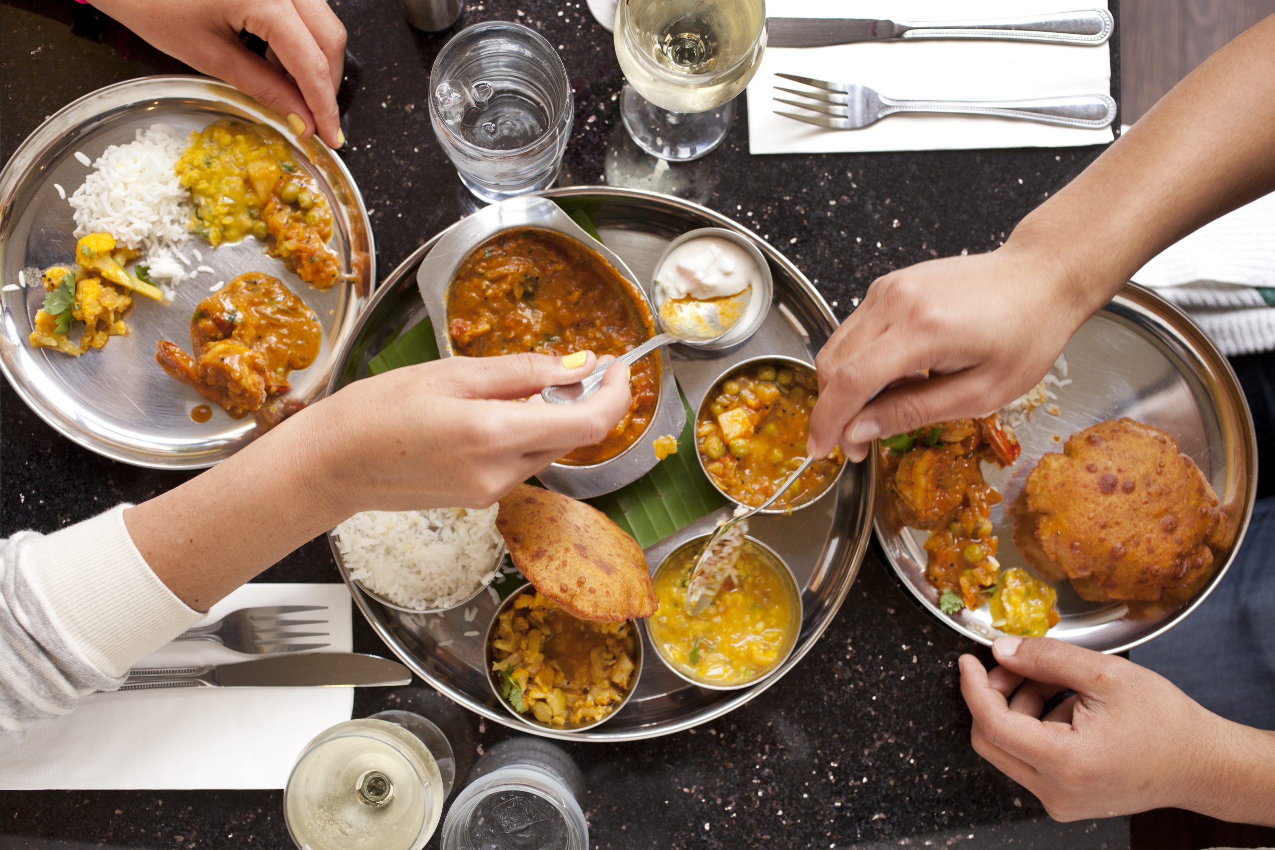 Two people sharing Indian food on a date.  An overhead shot of their hands reaching across the table and serving themselves the different dishes.